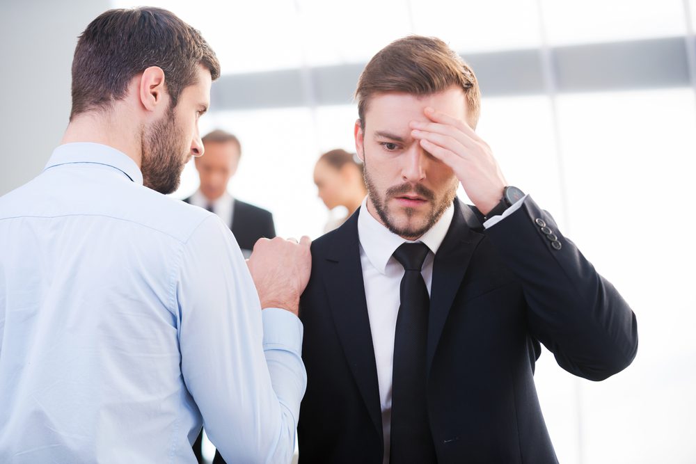 Bad news. Rear view of young businessman consoling his depressed colleague and holding hand on his shoulder with people standing in the background